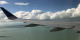 Photo of a plane's wing flying over the water with clouds in the background.